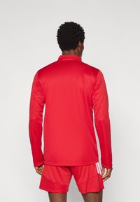 Κόκκινο αθλητικό μπλουζάκι με μακριά μανίκια και ψηλό γιακά, κατασκευασμένο από λείο ύφασμα, συνδυασμένο με κόκκινα σορτς με λευκές πλευρικές ρίγες.