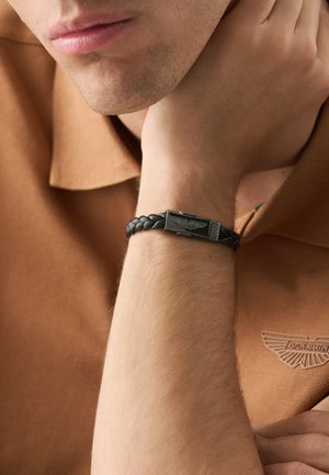 Čierny pletený kožený náramok s kovovou prackou, s detailom loga. V pozadí je svetlohnedé polo tričko s textúrou tkaniny.