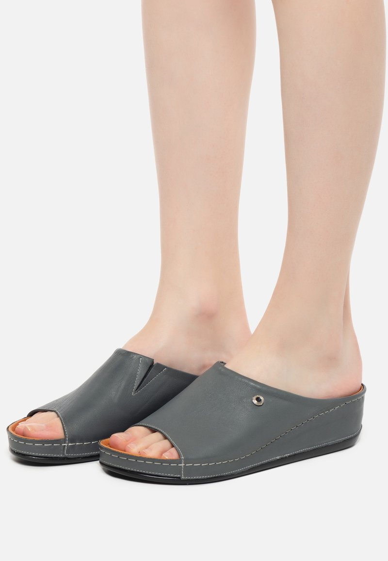 Pieds portant des chaussures gris foncé à enfiler, ouvertes à l’avant, avec des semelles compensées basses et des détails de coutures blanches visibles.