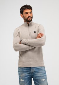 Hellbeige Zip-Jacke mit strukturiertem Muster, gerippten Bündchen und Saum, mit einem kleinen schwarzen Logo auf der Brust, kombiniert mit blauen, verwaschenen Jeans.
