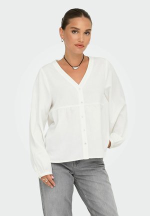 White, long-sleeve button-up blouse with a V-neck, textured fabric, and gathered waistline; worn with gray jeans and a black necklace.