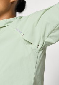 Chaqueta de color verde claro, texturizada, con un patrón de rejilla, que presenta un cuello alto y acentos de costura únicos cerca del hombro y las mangas.