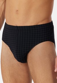 Torse masculin et cuisses supérieures portant un slip noir avec un motif de grille bleu clair subtil sur un fond uni.