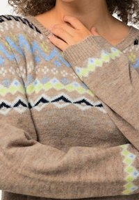 Nahaufnahme einer Person, die einen beigen Pullover mit blauen, grünen, schwarzen und weißen geometrischen Mustern trägt, mit einer Hand, die auf der Oberseite der Brust nahe dem Ausschnitt ruht.