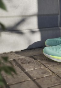 Scarpe da corsa azzurro chiaro e giallo neon poggiate su un pavimento in pietra contro un muro grigio illuminato dal sole con ombre.