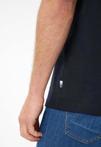 Zwarte katoenen t-shirt met korte mouwen, zijtag met blauwe en witte strepen, gedragen met blauwe jeans. Hand zichtbaar aan de zijkant.