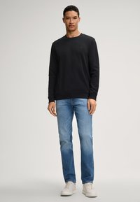 Schwarzes Sweatshirt mit Rundhalsausschnitt, kombiniert mit hellblauen Jeans und weißen Sneakern. Der Stoff wirkt weich, hat ein schlichtes Design und keine sichtbaren Muster.