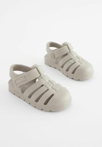 Beige Gummisandalen mit offenem Zehen- und Fersenbereich, die mehrere vertikale Aussparungen und einen verstellbaren Klettverschluss aufweisen. Strukturierte Sohle für besseren Grip.