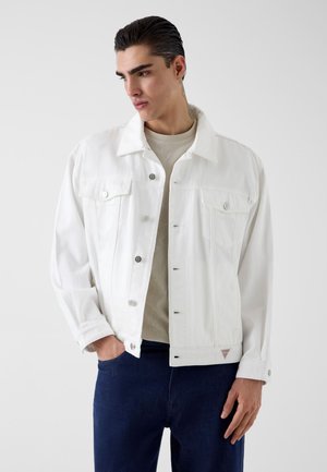 Giovane uomo che indossa una giacca di jeans bianca sopra una camicia beige e jeans blu scuro, in piedi con una mano in tasca, guarda a sinistra.