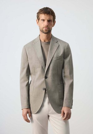 Hombre con pelo corto y barba que lleva una blazer beige de cuadros, camisa topo y pantalones de color claro, de pie frente a un fondo liso.