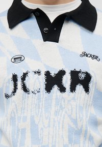 Κοντινή λήψη ενός πουλόβερ με μπλε και λευκό μοτίβο διαμαντιού, με μαύρο γιακά και τη λέξη "JACKR" πλεγμένη στο στήθος.