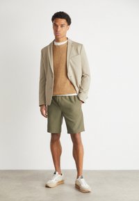 Beige blazer over brown knit sweater, olive shorts, and white sneakers with a gum sole, standing on a neutral floor against a light background.