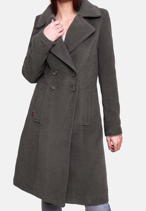 Femme portant un manteau en laine gris foncé à double boutonnage avec de larges revers et des poches latérales, tenant le col d'une main.