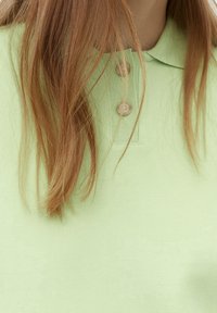 Jasnozielona koszulka polo wykonana z fakturowanej tkaniny, z kołnierzykiem i zapięciem na dwa guziki. Włosy częściowo zasłaniają dekolt koszulki.