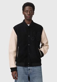 Ung mand iført en sort varsity-jakke med beige læderærmer, broderede "AS"-bogstaver og blå jeans, stående mod en ensfarvet baggrund.