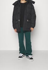 Chaqueta de plumas negra con capucha, cierre frontal con cremallera y dos bolsillos laterales, combinada con pantalones verde de pierna ancha y zapatillas negras y blancas.