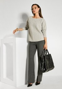 Vrouw in een beige gestructureerde trui, donkergrijze broek, zwarte hakken, met een grote zwarte handtas, staande naast een witte geometrische piedestal.