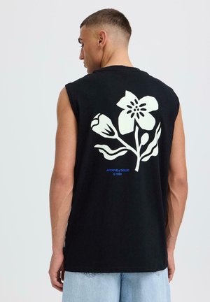 Homme portant un débardeur noir avec un grand motif de fleur blanche et le texte bleu "ARCHIVE of SOLID © 1999" au dos, assorti à un jean clair.