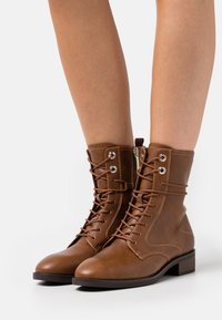 Braune Leder-Stiefeletten mit runder Zehenpartie, Schnürung vorne, silbernen Ösen und einem niedrigen, gestapelten Absatz. Glatte Textur mit Nähdetails.