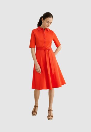 Mujer con un vestido naranja brillante hasta la rodilla con botones, lazo en la cintura, mangas hasta el codo y sandalias de cuña marrones, de pie frente a un fondo liso.