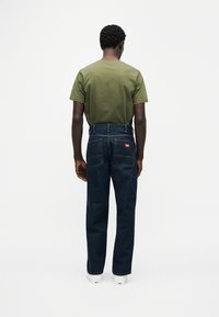 Λαδί πράσινο t-shirt σε συνδυασμό με σκούρο μπλε τζιν παντελόνι με ίσια γραμμή στα πόδια, και λευκά αθλητικά παπούτσια ορατά στο κάτω μέρος. Κόκκινη ετικέτα στο πίσω μέρος.