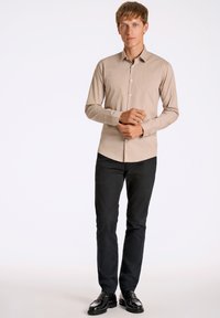 Camicia button-up di un beige chiaro con colletto a punta, design slim fit, abbinata a pantaloni neri e scarpe lucide nere.