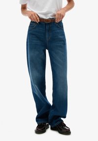 Blauwe denim jeans met een hoge taille, ruime pijpen en een gevlochten bruine riem. De stof heeft een gladde textuur en beschikt over klassieke zakken.