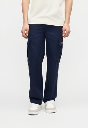 Άνδρας που φοράει ναυτικό μπλε παντελόνι cargo με τσέπες με καπάκι και λευκά αθλητικά παπούτσια, στέκεται μπροστά σε ένα απλό φωτεινό φόντο.