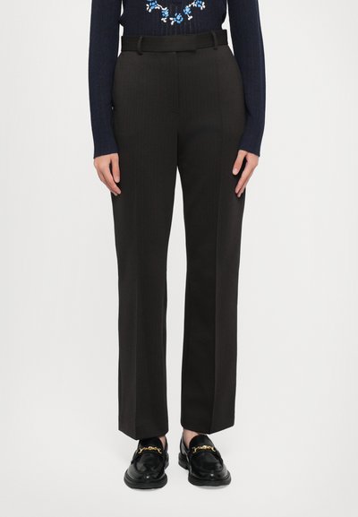 Melni pieguļoši bikses ar taisnu kāju, ar jostu cilpiņām un svītrām; valkātas ar melniem loafers un tumši zilu džemperi.