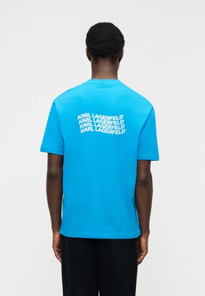 Persona che indossa una maglietta di un azzurro brillante con la scritta "KARL LAGERFELD" ripetuta quattro volte sulla parte superiore della schiena, in piedi davanti a uno sfondo semplice.