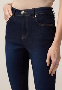 Jeans a vita alta di colore blu scuro realizzati in denim, caratterizzati da dettagli in metallo dorato, cuciture a contrasto e un classico design a cinque tasche.