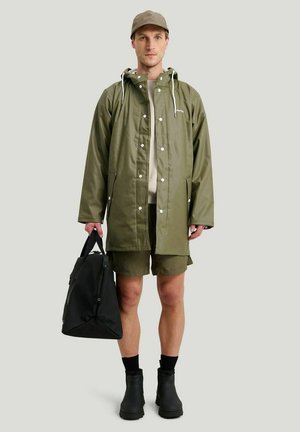 Olivgrön regnjacka med huva, tryckknappar och sidofickor; kombinerad med svart väska, beige skjorta, gröna shorts och svarta stövlar.