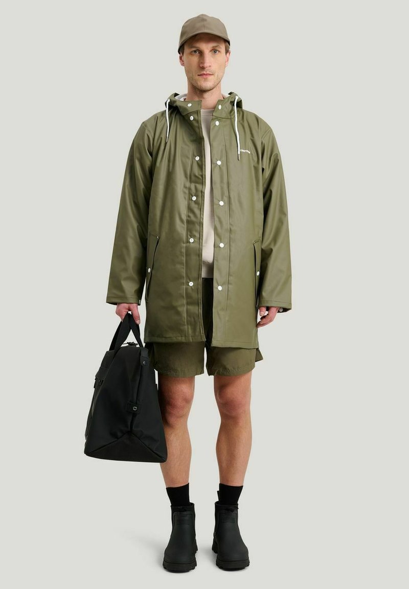 Olivgrön regnjacka med huva, tryckknappar och sidofickor; kombinerad med svart väska, beige skjorta, gröna shorts och svarta stövlar.