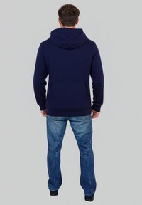 Marineblaue Kapuzenjacke aus mittelschwerem Stoff, mit einer großen Kapuze, Fronttasche und gerippten Bündchen. Kombiniert mit blauen Jeans.