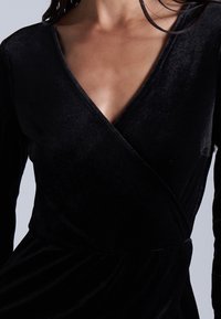 Vestido de terciopelo negro con mangas largas, escote en V y diseño envolvente. Presenta una textura suave y un acabado liso.