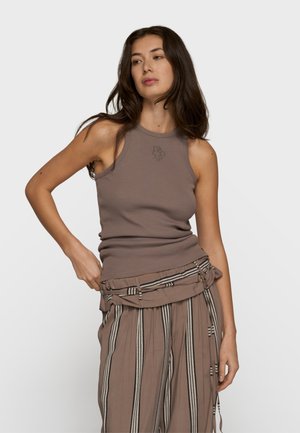 Frau mit langem dunklem Haar, die ein ärmelloses taupefarbenes Oberteil und lockere, gestreifte taupefarbene Hose mit passendem Gürtel trägt, steht vor einem schlichten Hintergrund.