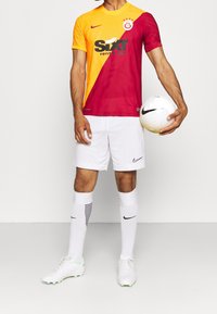 Camiseta local del Galatasaray en naranja y rojo con el logo del patrocinador en negro, combinada con pantalones cortos blancos, medias blancas y botas con detalles verdes, sosteniendo un balón de fútbol.