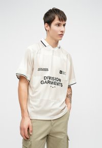 Lys beige sportstrøje med sorte accenter; har et tekstureret ternet mønster, kortærmede og trykte logoer i sort.