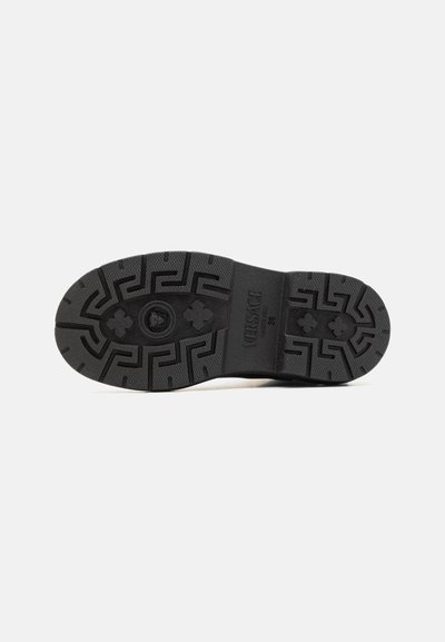 Suela de zapato de goma negra con un patrón texturizado, con un logo de Medusa en el centro y un diseño de banda de rodadura geométrico para tracción.