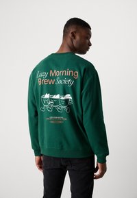 Sweat-shirt en polaire verte avec un graphisme ludique de trois tasses de café au dos. Le texte comprend "Société du café des matinées paresseuses" et une invitation.