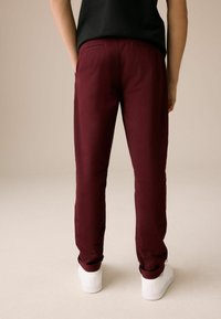 Bordeaux slim-fit bukser med en glat tekstur og to baglommer, parret med hvide sneakers, set bagfra mod en neutral baggrund.