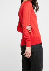 Maglione rosso a maglia con maniche lunghe, scollo a barca, polsini e orlo a coste. Abbinato a jeans scuri, che mostrano una texture liscia e una silhouette aderente.