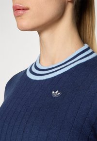 Donkerblauwe geribbelde sweater met een lichtblauwe gestreepte kraag en een klein geborduurd logo op de borst. Close-up van de halslijn.