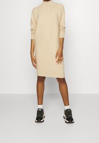 Abito a maglia beige con collo alto, maniche lunghe e polsini a coste, abbinato a sneaker bianche e nere e calzini bianchi alla caviglia.