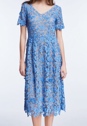 Femme portant une robe bleue en dentelle à manches courtes et longueur genoux, décorée de motifs floraux avec un décolleté en V.