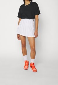 T-shirt cropped nera, gonna plissettata bianca, sneaker arancioni alte e calze bianche. La gonna presenta un bottone decorativo nero e una fascia in vita decorativa.