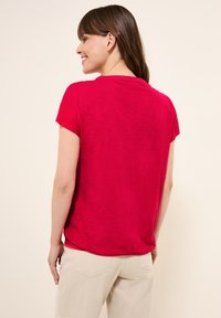 Haut rouge à manches courtes avec une coupe décontractée et un col rond. Tissu léger et texturé, associé à un pantalon de couleur claire. Vue de dos.