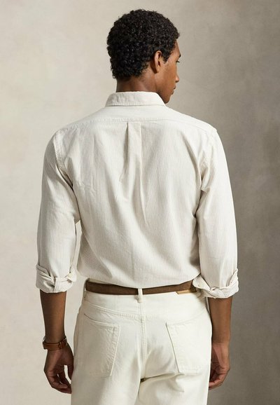 Homme aux cheveux bouclés portant une chemise à manches longues rayée clair et un pantalon crème, vu de dos, manches retroussées et ceinture marron.