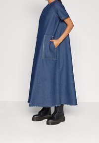 Denimmekko, täyspitkä, sininen, lyhyet hihat, iso etutasku, raaka helma, yhdistetty mustiin nauhakengiin. Pehmeä pinta.
