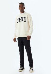 Sudadera de crema con "OHIO" en letras negras en negrita, combinada con pantalones negros y zapatillas negras con suelas blancas, de pie sobre un fondo blanco.